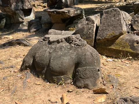       Weather-worn torso of a stone statue lying broken among boulders and dry grass in temple grounds
  
