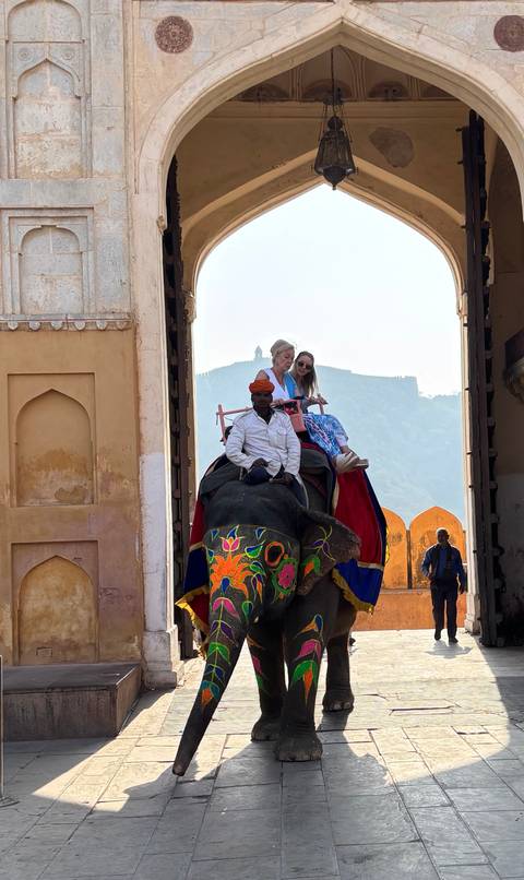       Decorated elephant carrying two tourists passes through a grand fort gateway
  