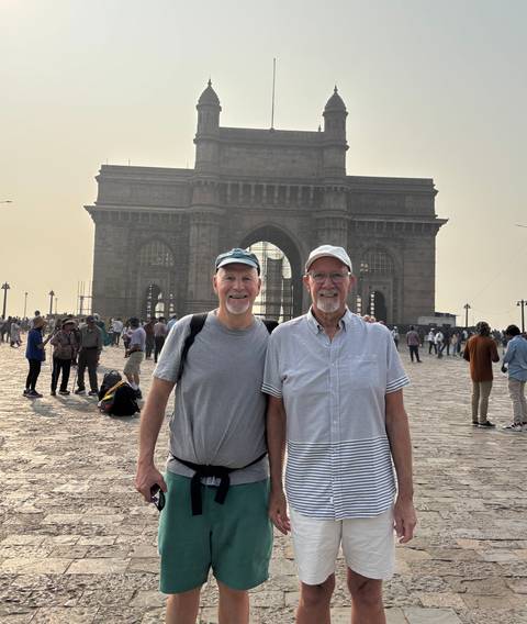      Travel duo stands before the imposing Gateway of India with crowds behind them
  