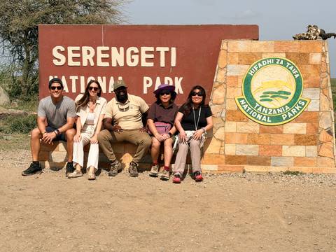       Travellers pose beside the Serengeti National Park entrance sign on a sunny day.
  
