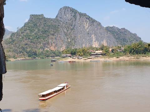       Riverboat cruises past dramatic limestone cliffs and riverside village in Laos.
  