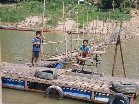       Two boys stand on a bamboo-and-metal floating footbridge over a calm river on a sunny day.
  