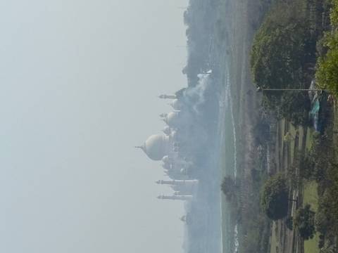       Distant, hazy view of the Taj Mahal beyond smoky fields under a pale sky.
  