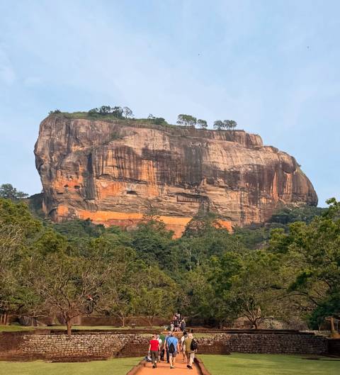       Sheer walls of Sigiriya Rock Fortress rise above lush Sri Lankan jungle under a blue sky
  