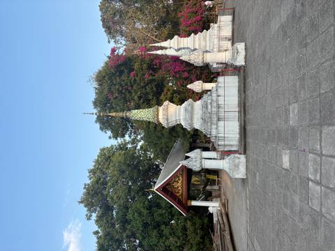      White stupa monuments and ornate pavilion surrounded by flowering trees in a temple courtyard.
  