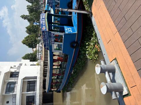       Blue riverboat docked beside riverside promenade with floating plants.
  