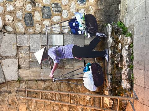       Street vendor wearing conical hat carrying balanced baskets of goods beside stone wall.
  