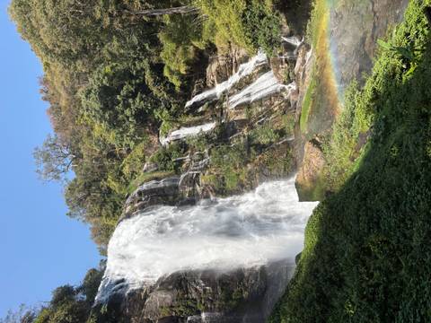       Tall waterfall plunging over rocky cliff in jungle with visible rainbow in mist.
  
