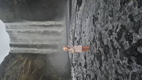       Person in winter coat standing on black gravel before the misty curtain of Skógafoss waterfall
  