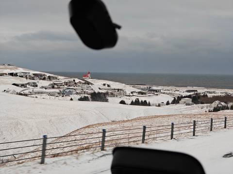       Snow-covered coastal village and church viewed from a vehicle window with ocean beyond
  