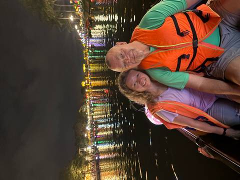       Smiling couple in life vests sit on a small boat at night with Hoi An’s colorful riverside lanterns reflected in the water behind them.
  