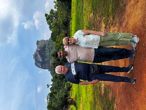       Three smiling travelers posing arm-in-arm on a grassy clearing with a steep rock monolith rising in the background.
  