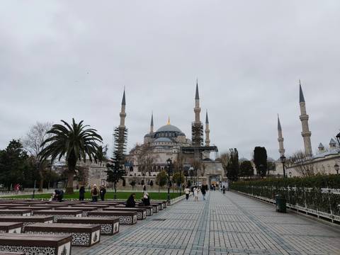       Visitors stroll in front of Istanbul’s Blue Mosque, partially covered by restoration scaffolding on a grey day.
  