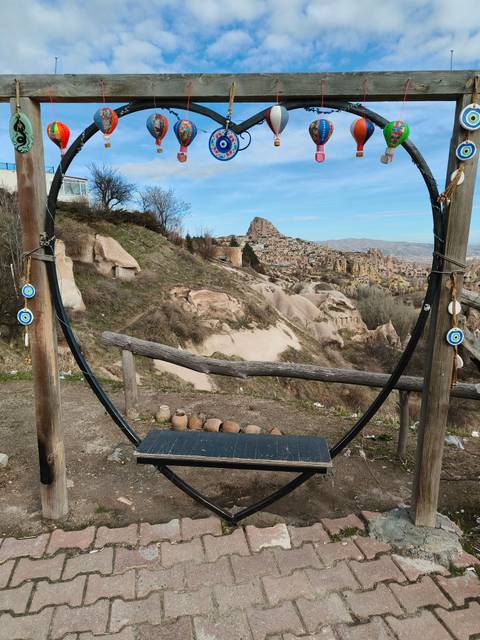       Scenic overlook with a large wooden swing frame decorated with Nazar evil-eye charms, looking out over Cappadocia’s rocky fairy-chimney landscape and Uchisar Castle.
  