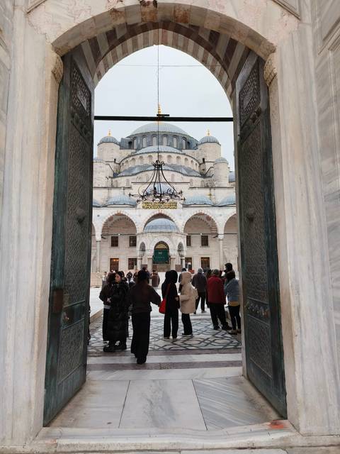       Visitors enter the grand marble courtyard of an Ottoman mosque through ornate bronze doors, framed by columns and arches.
  