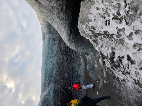       Traveller in red safety gear stands beside a translucent blue ice cave wall on a glacier.
  
