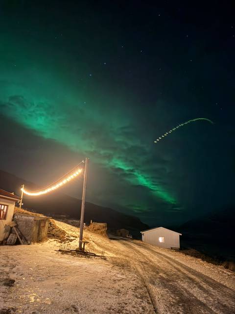      Green aurora borealis swirling above a valley with a string of festive lights and a cottage roof
  