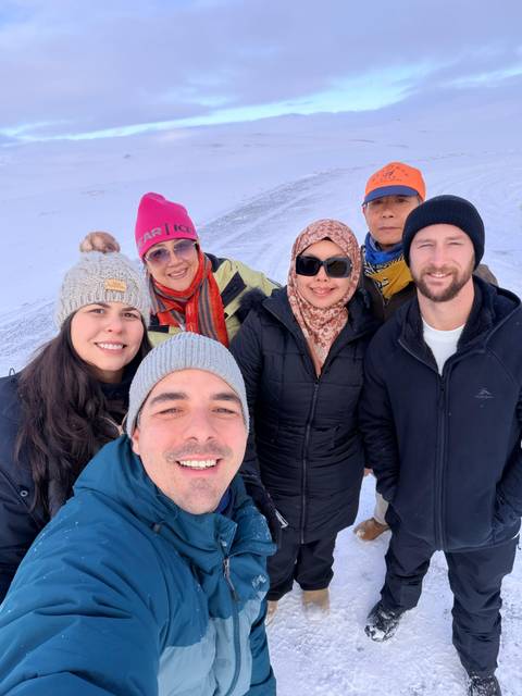       Smiling group selfie of six travellers in winter clothing on a snowy plain
  