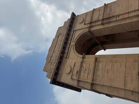       Low-angle view of the sandstone India Gate war memorial against a partly cloudy sky.
  