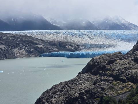      A massive blue glacier descends into a pale grey lake framed by rugged dark rock walls.
  