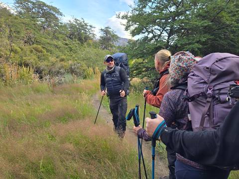       Hikers carrying backpacks and trekking poles chat while walking along a grassy trail lined with shrubs and trees.
  