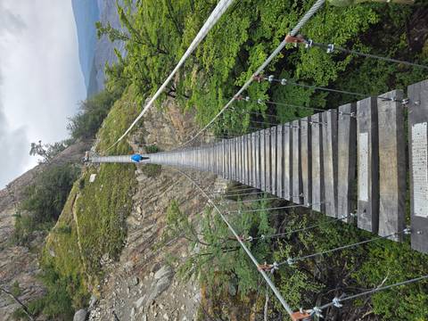       A long wooden suspension bridge stretches across a deep green ravine toward a lone hiker in a blue jacket.
  