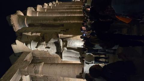       Crowds gather at night beneath towering statues and columns in Luxor Temple lit by warm spotlights.
  