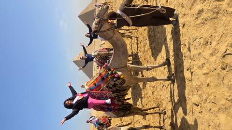       Travellers cheer while riding camels in the sandy desert with the pyramids in the background.
  