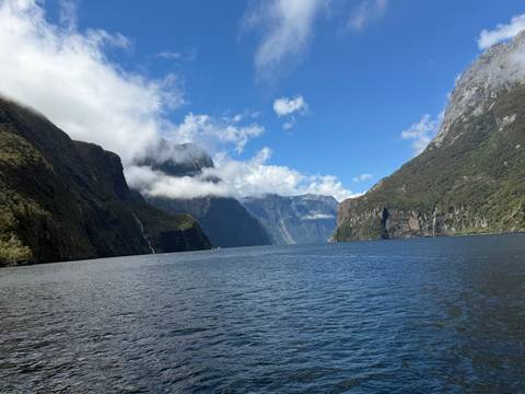       A dramatic fjord scene with steep forested cliffs, waterfalls, and mist drifting above deep blue water beneath a partly cloudy sky.
  
