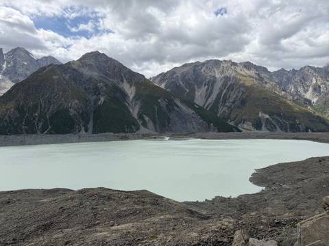       Pale glacial lake surrounded by towering rocky peaks and scree slopes under a cloudy sky.
  
