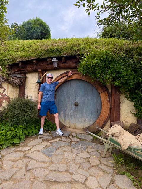       A visitor smiles beside the iconic round wooden door of a Hobbiton house set in lush greenery.
  