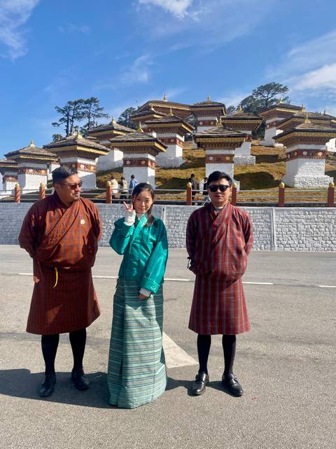       Three travelers in traditional dress pose in front of the white and gold chortens of Dochula Pass on a sunny day.
  