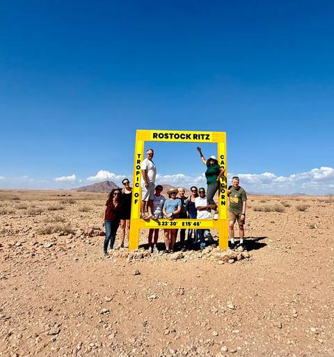       Travel group poses around a bright yellow Tropic of Capricorn frame in a vast Namib desert landscape.
  
