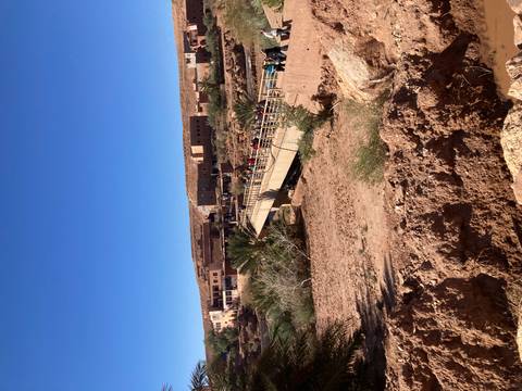       Bridge spanning a dry riverbed leading to adobe buildings under bright blue Moroccan sky.
  