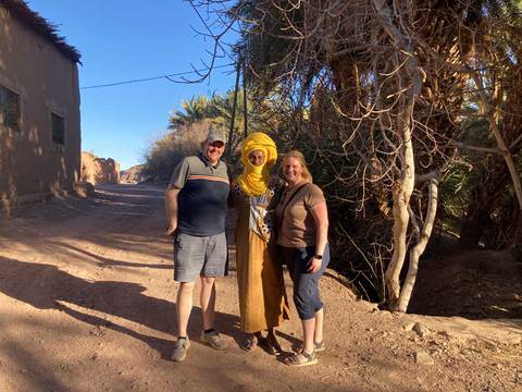       Two tourists pose with a local man in yellow turban on a dusty palm-lined village track.
  