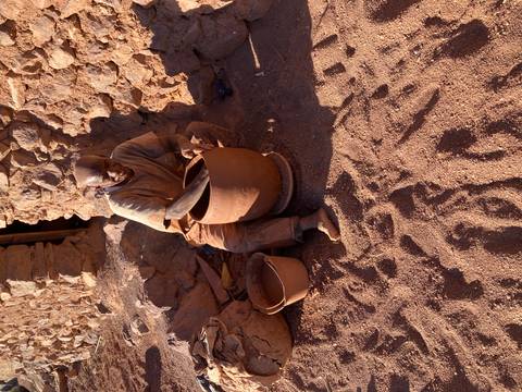       A potter works a clay vessel with bare feet in the sand beside a stone wall.
  