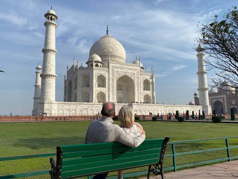       Couple sits arm-in-arm on a bench gazing at the Taj Mahal across green lawns under a clear blue sky.
  