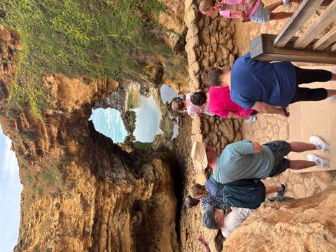       Travellers descend stairs toward a natural rock arch and tidal pool set in golden cliffs.
  