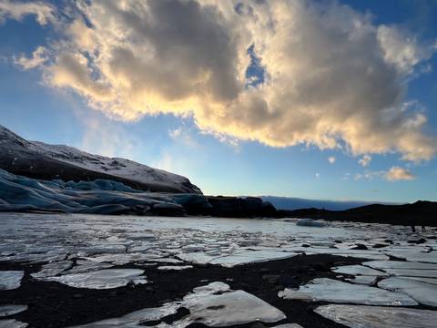       Frozen lagoon with scattered blue icebergs beneath snowy mountains and a dramatic sky.
  