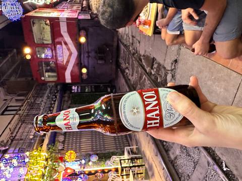       Hand holding a Hanoi beer bottle with a red locomotive approaching on train street at night.
  