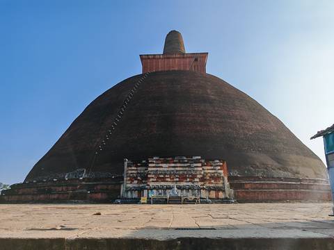       Massive brick stupa rising toward a clear blue sky with a small entrance base, photographed from ground level.
  