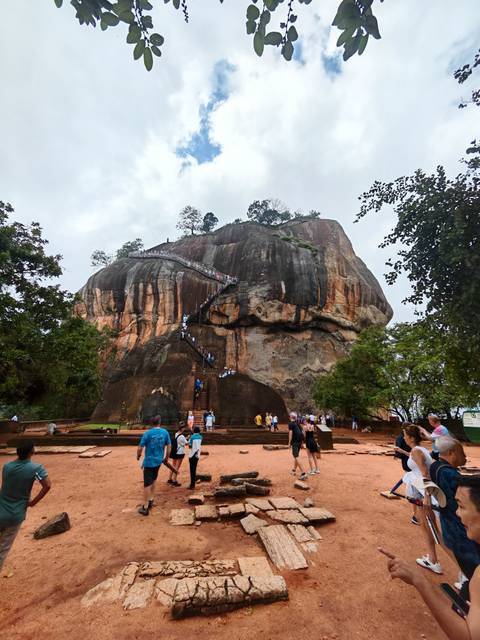       Visitors climb a steep staircase up the iconic Sigiriya rock fortress, its streaked cliffs towering over surrounding greenery.
  