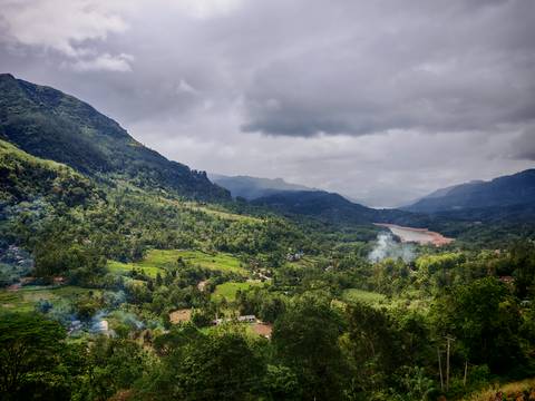       Expansive view of a lush green valley with winding river and layered mountains under a moody cloudy sky.
  