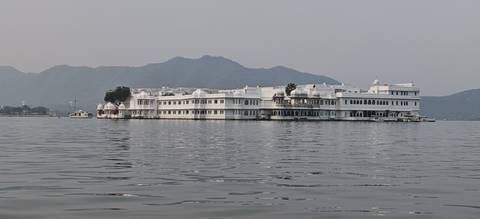       The white Lake Palace floats on calm waters with hazy hills in the background.
  