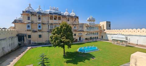       Historic palace courtyard with manicured lawn and turquoise fountain under blue sky.
  