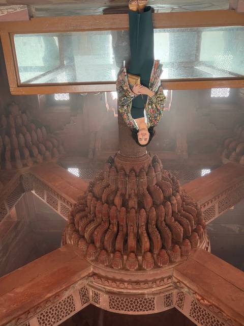      Woman stands beneath an elaborate carved sandstone pillar inside Fatehpur Sikri.
  
