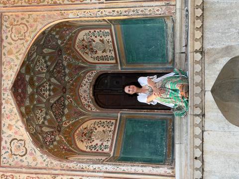       Woman sits in a richly painted Mughal archway surrounded by floral murals.
  