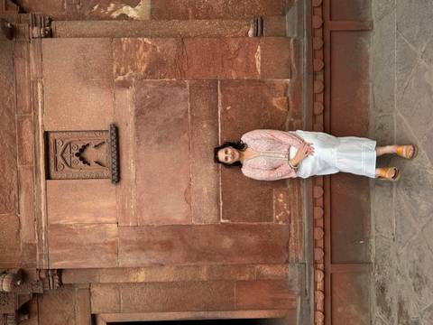       Woman sits against a weathered red sandstone wall with minimal carvings.
  