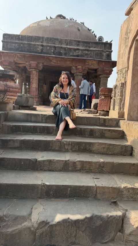       A barefoot woman sits casually on worn stone steps of an old sandstone temple or fort.
  