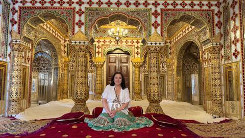       Woman sits inside a dazzling mirrored and gilded chamber adorned with intricate Rajasthani decor.
  
